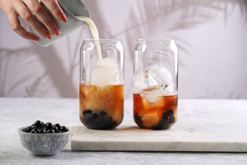 Female hand pouring milk into glass with black tea, ice cubes and cooked tapioca pearls for trendy...