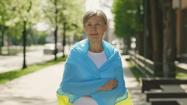 Mature woman carries a the ukrainian flag standing on the street looking at the camera on the blurred background. People portrait. Political concept