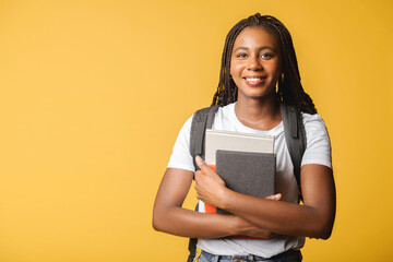 Happy excited female student with backpack standing isolated on yellow and holding books and...