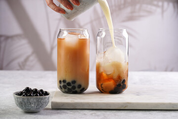 Female hand pouring milk into glass with black tea, ice cubes and cooked tapioca pearls for trendy bubble boba ice tea, two small grey ceramic bowls on marble board on pastel tropical background