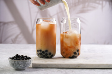 Female hand pouring milk into glass with black tea, ice cubes and cooked tapioca pearls for trendy...