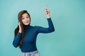 Surprised young beautiful girl with long hair in blue sweater, blue jeans is standing on turquoise background pointing by index finger up with surprised eyes looking at camera with empty space  for ad