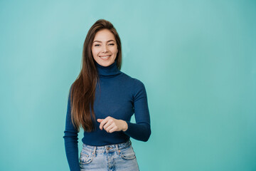 Young successful Italian girl with long loose hair in a blue sweater and jeans, standing on a turquoise background in the studio, broad smiling, looking at camera, happy to be active and productive.