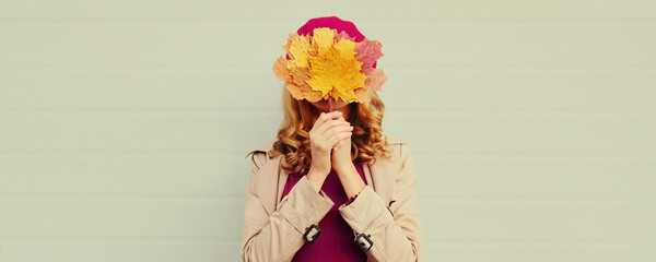 Autumn portrait of happy woman covering her eyes with yellow maple leaves on gray background