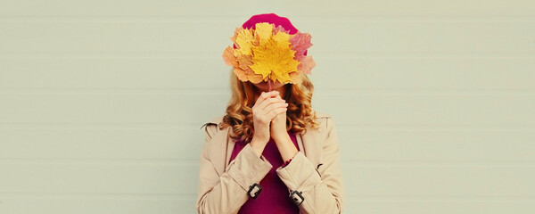 Fototapeta premium Autumn portrait of happy woman covering her eyes with yellow maple leaves on gray background