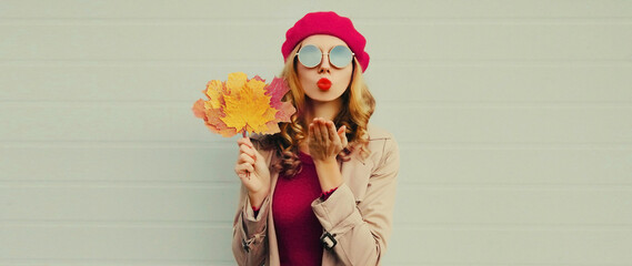 Autumn portrait of beautiful young woman model with yellow maple leaves blowing her lips with lipstick wearing pink french beret on gray background