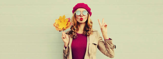 Autumn portrait of beautiful young woman model with yellow maple leaves blowing her lips with lipstick wearing pink french beret on gray background