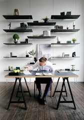 A young man is sitting at his desk. He is dressed in a white plane and classic plaid trousers. There are many shelves in the background. The concept of busy hardworking people in the office