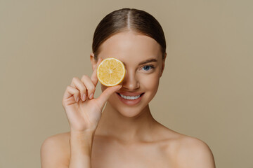 Portrait of good looking woman has dark combed hair perfect healthy skin eats organic food covers eye with slice of lemon stands bare shoulders indoor smiles toothily isolated over beige background