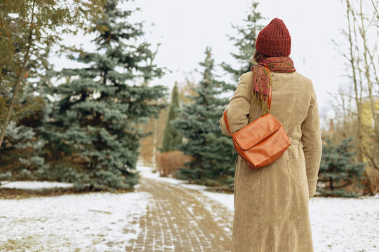 Back view of woman in warm coat and hat walking on snowy pathway in winter park in city 