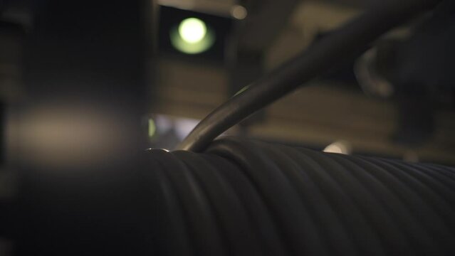 The cable slowly unwinds from a large spool at the cable factory. Close-up. A bright lamp in the background shines brightly. Production process at the cable factory. 4K
