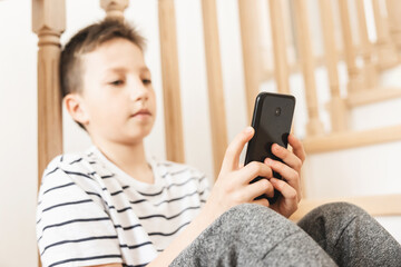 Teenage boy sitting on step of stairs using smartphone and playing video games on internet online at cozy home. Child holding mobile phone and looking at screen. Social media