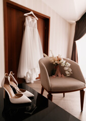 An elegant wedding dress hangs by a door. Nearby are women's shoes and bouquet