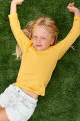 Portrait cute blonde girl with closed eyes lying, stretching on green grass. Mock up yellow t shirt. Top view.
