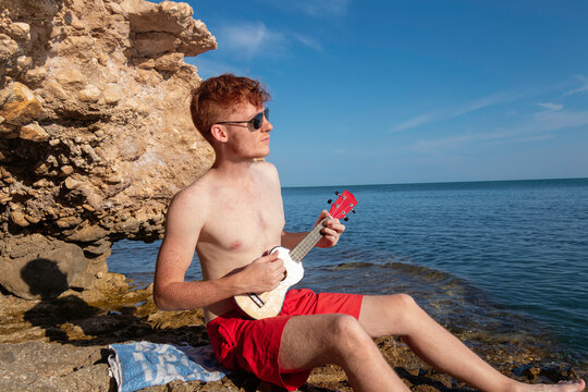 young red hair caucasian man playing the ukelele on the beach in sunny day
