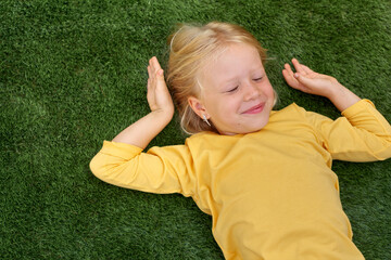 Portrait cute blonde girl with closed eyes lying on green grass. Mock up yellow t shirt. Top view.