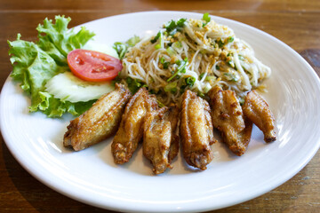 Fried wing chicken served with spicy rice flour noodle salad and vegetables; cucumber, tomato, and green lettuce from Thailand.