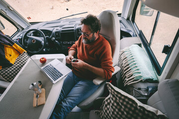 Digital nomad and travel wanderlust lifestyle people concept. One man viewed from above working on laptop and drinking coffee inside a modern camper van. Job and vacation lifestyle. Holiday and work - obrazy, fototapety, plakaty
