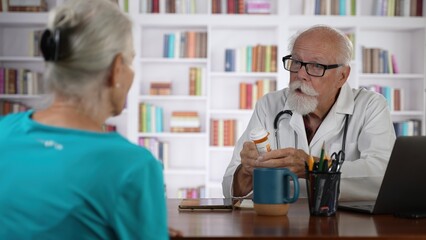Fototapeta na wymiar Mature senior male doctor discussing treatment with prescription drug to female patient in modern clinic with books background.