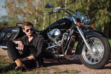 Handsome male biker is sitting on the ground with his motor bike outdoors