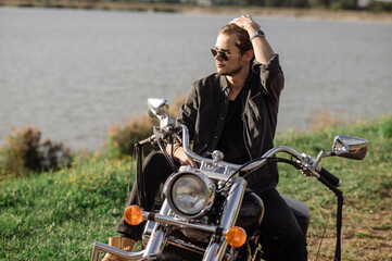 Fototapeta na wymiar Young handsome male biker is sitting on his motor bike outdoors at the lake background
