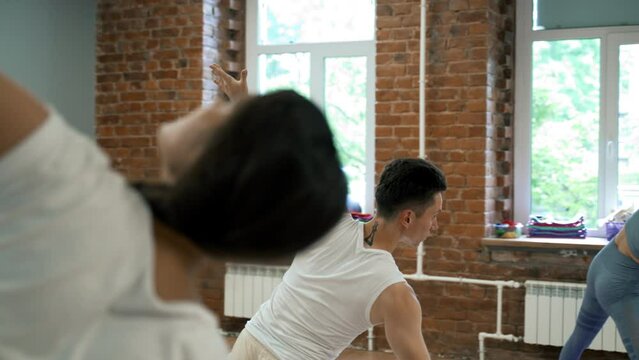 People lean to side spreading hands at yoga class in studio with bright windows. Coach teaches women to do yoga pose Triangle closeup slow motion