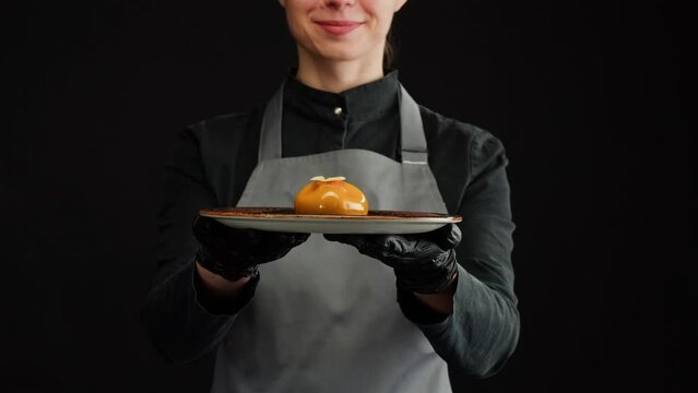 Woman chef holding mousse orange cake, creamy dessert close-up. Almond dacquoise, raspberry confit, crispy layer with caramelized hazelnuts and raspberry powder, mousse based on bitter chocolate.