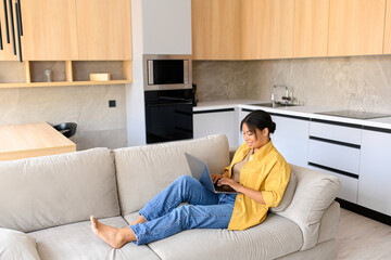 Relaxed young African-American female freelancer sitting on the couch watching movie series on laptop or working remotely, typing emails, side view