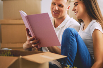 Cute couple unpacking cardboard boxes in their new home, sitting on the floor and looking at a family album
