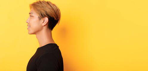 Profile of stylish handsome asian guy with fair hair looking left and smiling, standing over yellow...