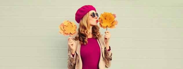 Autumn portrait of beautiful young woman model with yellow maple leaves blowing her lips with lipstick wearing pink french beret on gray background