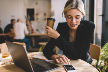 Millennial female with modern laptop checking received email message on smartphone device during coffee break for remote working, skilled woman in earbuds choosing music playlist on cellphone