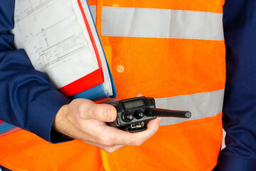 The torso of a man in an orange reflective vest with building plans in a folder, holding a...