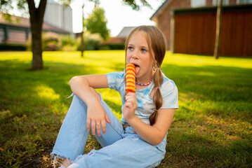 A beautiful European girl eats bright ice cream on a background of green grass. Girl with freckles on the background of a green lawn. Redhead girl with ice cream. Child on vacation. Happy  children 