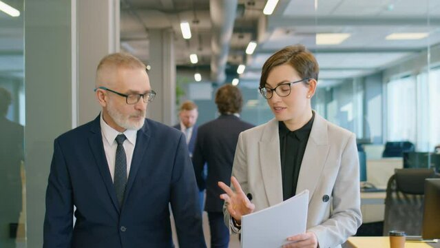Waist up shot of young businesswoman explaining documents to senior male colleague while walking together through office