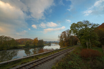 Fototapeta na wymiar Eisenbahnschienen an einem Fluss mit Wald im Herbst bei blauem Himmel mit Wolken 