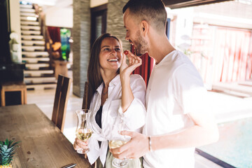 Caucasian wife feed husband enjoying time for eating togetherness during brunch at home interior, cheerful woman with glass of white wine holding cherry tomato during positive date with boyfriend
