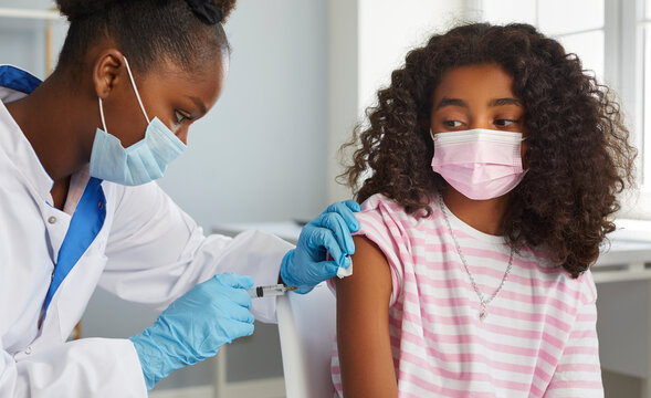 Vaccinations for children. Brave African American teenage girl receives booster dose of coronavirus vaccine. Female nurse in mask and gloves gives injection to child's hand in clinic. Medical banner.