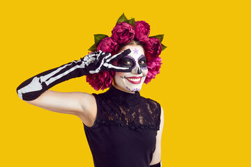 Happy beautiful woman with skull face makeup and flowers on head does peace victory sign with two...