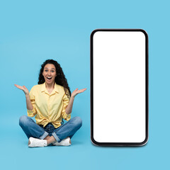 Excited young woman sitting near big cellphone with empty space for mobile app or website on screen, mockup