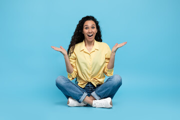 Puzzled excited young woman shrugging shoulders and smiling at camera on blue studio background, full length