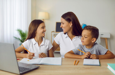 Happy family doing homework. Responsible parent helping children get ready for school project. Daughter and son together with mother sitting at desk, writing and drawing, using laptop and having fun