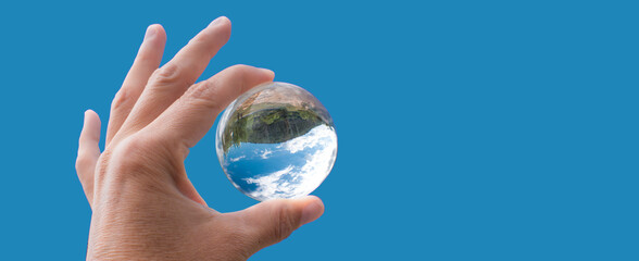 a hand is holding a glass sphere in which a landscape and blue sky with clouds is seeing, blue background