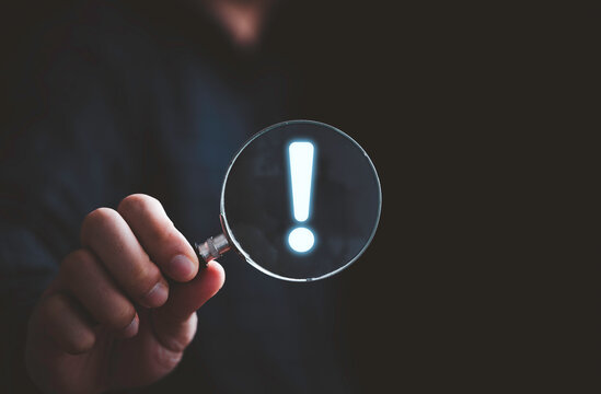 Hand holding magnifier glass with red triangle caution warning sign for notification error and maintenance concept.