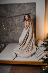 A young woman wrapped in a blanket is standing on a bed near a Christmas tree.