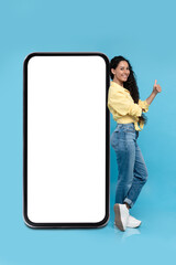 Lovely young woman standing near big mobile phone with empty white screen and showing thumb up gesture, mockup