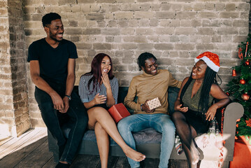 Diverse group of cheerful friends sitting on the couch by Christmas tree and opening gifts together