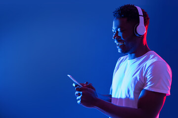 Happy millennial black guy in wireless headphones using smartphone in blue neon light, copy space