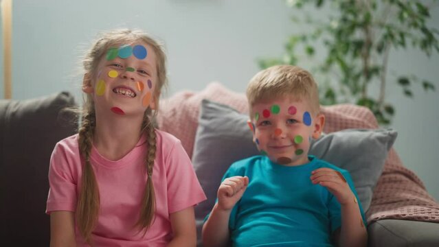 Joyful elder sister and toddler brother with colorful dot stickers on faces sit on sofa slow motion. Children holiday celebration. Facial decoration