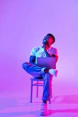 Thoughtful young black guy sitting on chair with modern laptop, looking at empty space above his...